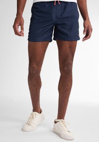 Costume de baie bleu marin cu cordon la talie și buzunare laterale. Fabricat dintr-un material ușor, având o etichetă de marcă la tiv.