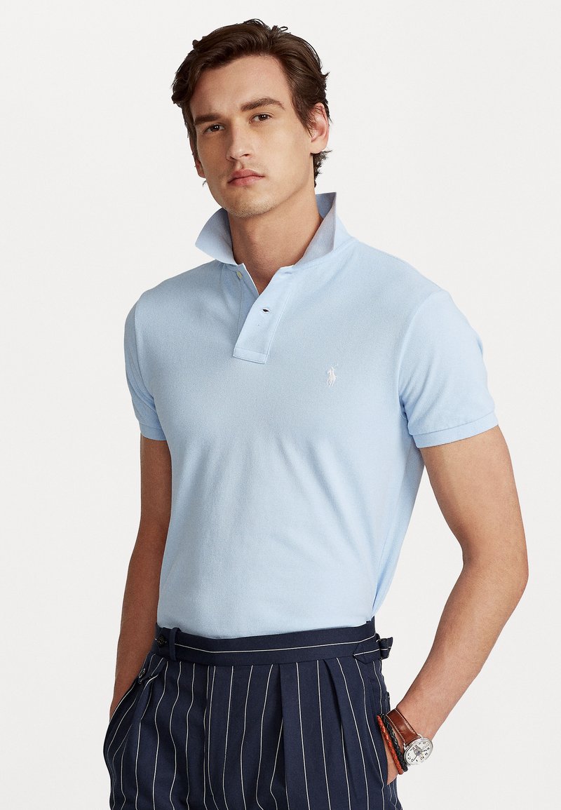 Light blue polo shirt with short sleeves, collar, and subtle logo. Material appears soft with a textured finish. Paired with striped navy trousers.