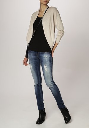 Žena v béžovém cardiganu, černém topu, opotřebovaných modrých úzkých džínách, černých kotníkových botách a s černou kabelkou přes rameno, stojící s rukou v bok.