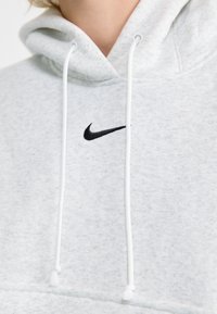 Svijetlosiva kapuljača s mekom teksturom, s crnim Nike logotipom na prsima, bijelim vezicama i kengur džepom.