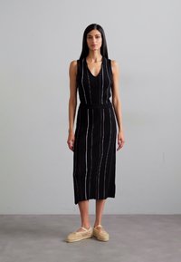 Rochie neagră fără mâneci cu dungi verticale albe, decolteu în V și siluetă ajustată, asortată cu pantofi slip-on din material țesut de culoare deschisă.
