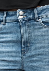 Κοντινό πλάνο από ανοιχτό μπλε τζιν τζιν με διπλά μεταλλικά κουμπιά και ορατή ραφή στη μέση ενός ατόμου που φορά μαύρο τοπ.