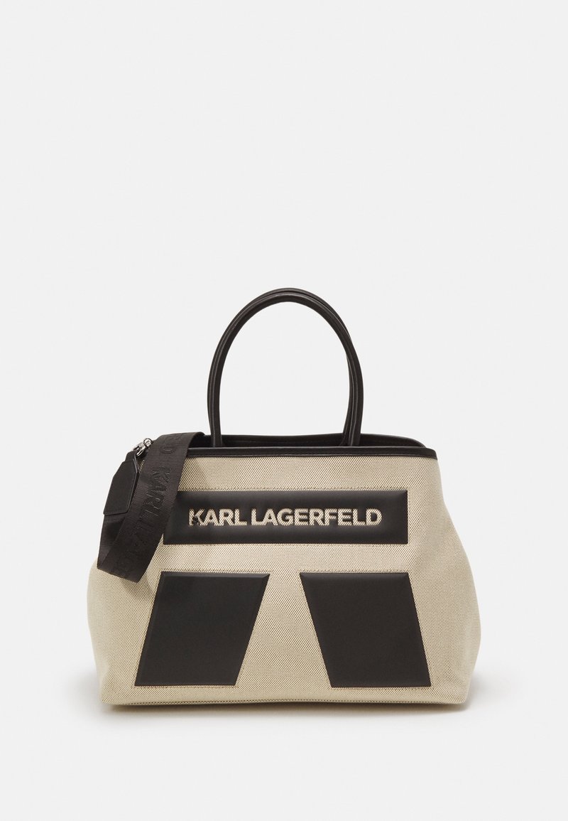 Sac à main en toile beige avec accents en cuir noir, double poignée et patch logo. Présente une forme structurée et une fermeture éclair.