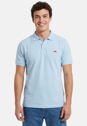 Jeune homme souriant portant un polo bleu clair avec un petit logo sur la poitrine gauche et un jean bleu sur fond blanc.