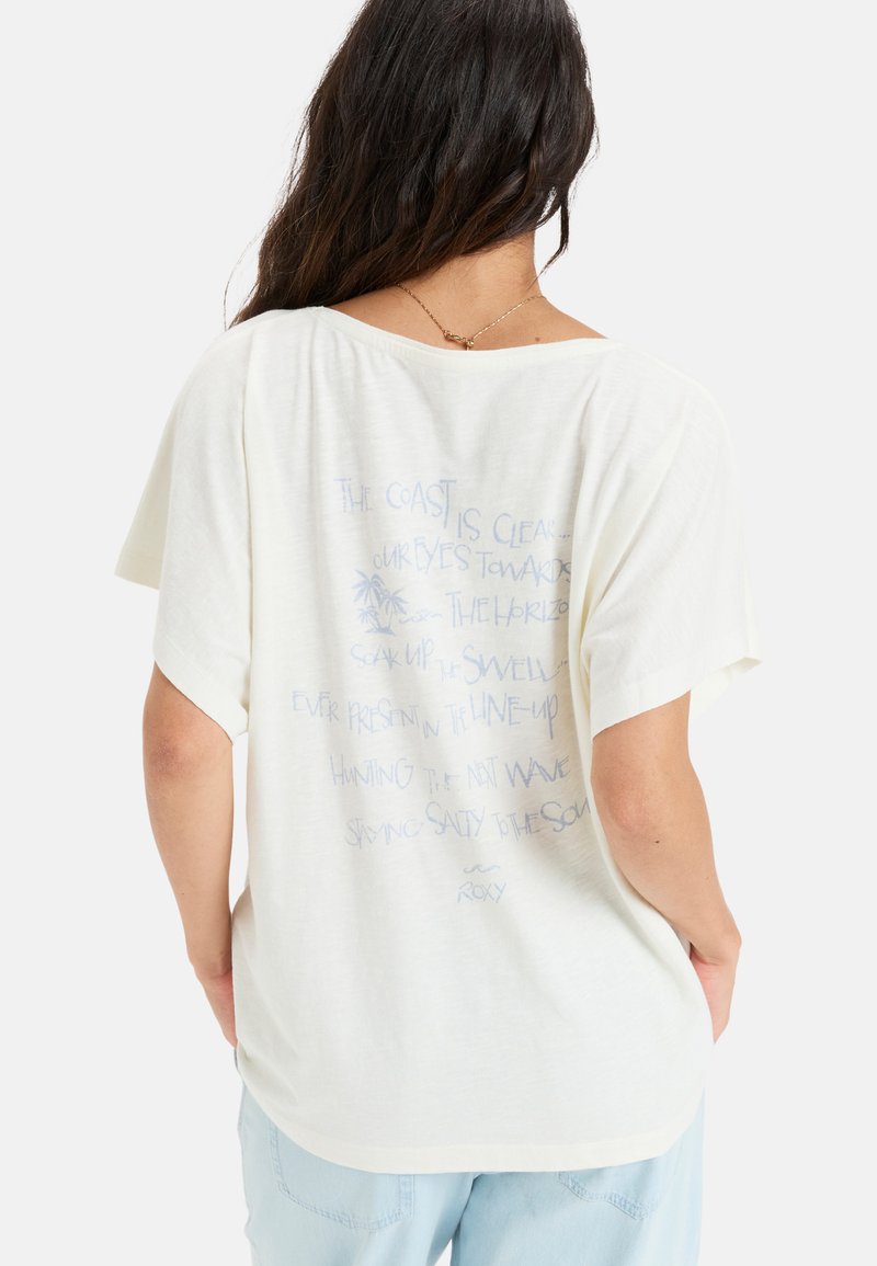 Camiseta oversized de color crema claro con mangas cortas. La parte posterior presenta un diseño de texto azul con frases temáticas de surf y una pequeña ilustración de una palmera.
