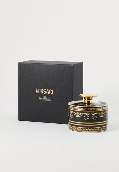 Świeca Versace o okrągłym, czarnym ceramicznym korpusie, ze złotymi akcentami i misternymi wzorami. Towarzyszy jej czarne pudełko z złotym logo.