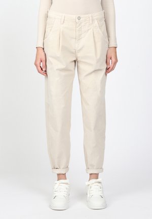 Pantalon beige plissé à taille haute avec revers roulés, porté avec des baskets blanches et un haut beige à manches longues, sur un fond uni.