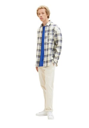Plaid button-up shirt in beige with navy and black accents, layered over a blue t-shirt; paired with light beige pants and white sneakers.