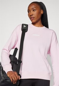 Sudadera rosa claro de manga larga y cuello redondo, con el logo blanco de Adidas en el pecho. Bolsa negra con una correa.
