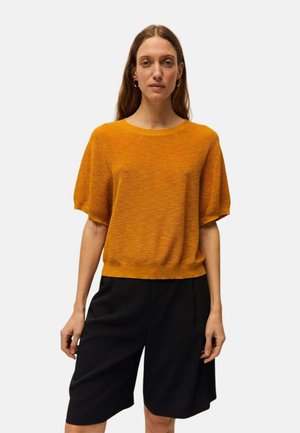 Mujer con un suéter naranja suelto de manga corta combinado con pantalones cortos negros hechos a medida hasta la rodilla sobre un fondo liso.