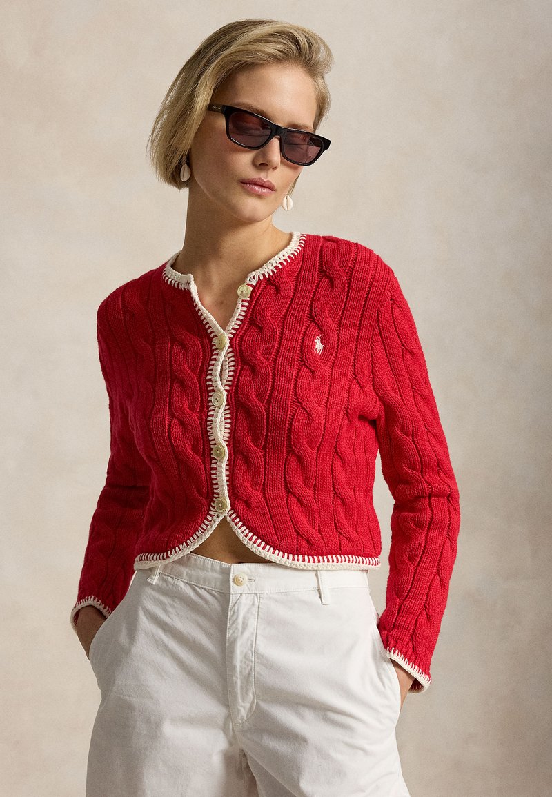 Kobieta w czarnych okularach przeciwsłonecznych, czerwonym kardiganie na guziki z białym obszyciem i białych spodniach z wysokim stanem, ręce w kieszeniach.