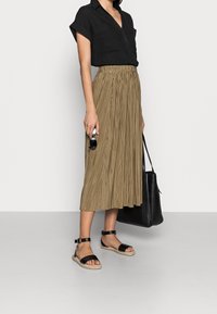 Svart kortärmad skjorta med knappar, plisserad khaki midikjol, svarta espadrillosandaler, håller i solglasögon och har en svart tote-väska.