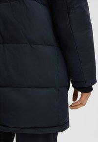 Donker marineblauwe pufferjas met horizontale gestikte vulling, een hoge kraag en een aansluitende polsmanchet. Glad textuur met een strakke vormgeving.