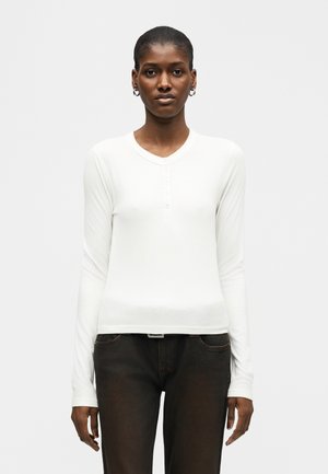 Γυναίκα με εφαρμοστό λευκό μπλούζα μακρυμάνικο henley και σκούρο παντελόνι, στέκεται μπροστά σε απλό ανοιχτό γκρι φόντο.