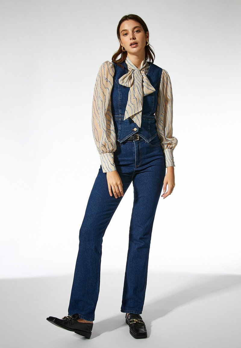 Overoles de mezclilla en azul oscuro con un diseño ajustado, combinados con una blusa de color beige claro con patrones de olas y un gran lazo en el escote.