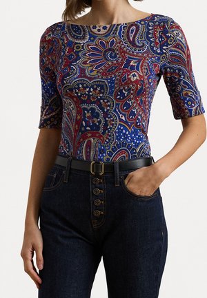 Frau trägt ein enganliegendes, paisleygemustertes Oberteil in Rot, Blau und Gelb, kombiniert mit hoch taillierten dunklen Jeans und einem schwarzen Gürtel.