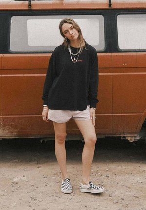 Kobieta stojąca z jedną nogą zgiętą, ubrana w czarną bluzę, jasne szorty i buty slip-on w kratę, na tle rdzawozłotego vana.