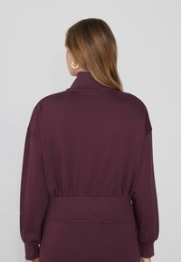 Maglia a collo alto borgogna con vita elasticizzata e maniche lunghe. Texture liscia con una silhouette aderente. Orecchini a cerchio dorati visibili.