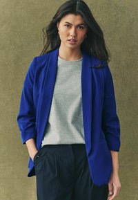 Royal blue blazer with structured shoulders, worn over a light gray t-shirt. The ensemble features relaxed-fit navy pinstriped trousers.