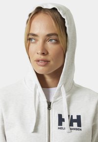 Sudadera con cremallera en gris claro que presenta un logo en la parte frontal, cordones en la capucha y un acabado de tela suave y texturizada.