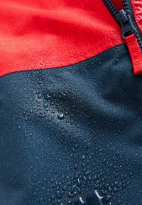 Veste imperméable présentant une partie supérieure rouge et une partie inférieure bleue marine, avec une surface texturée ornée de gouttes d'eau. Fermeture éclair visible.