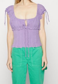 Top corto smock lilla con maniche a volant e chiusura a nodo sul collo, abbinato a pantaloni verdi a vita alta con tasche frontali.