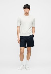 Jeune homme debout portant un haut en maille à manches courtes blanc, un short bleu marine, des chaussettes blanches montantes et des baskets blanches sur un fond uni.