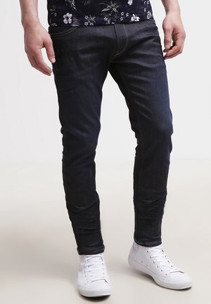 Pantalones vaqueros oscuros de corte ajustado, con diseño tradicional de cinco bolsillos y costuras destacadas, combinados con zapatillas blancas.
