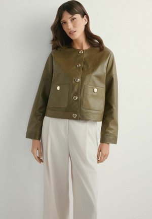 Kobieta w oliwkowo zielonej skórzanej kurtce zapinanej na guziki z dwoma kieszeniami z przodu i białych szerokich spodniach, stojąca na jednolitym tle.