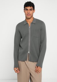 Mann som har på seg en grå jakke med glidelås og brystlomme, samt beige bukser, står mot en enkel hvit bakgrunn.