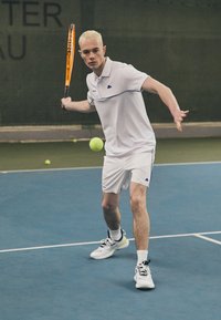 Weißes Polo-Shirt mit blauen Akzenten, weiße Shorts und Tennisschuhe. Hält einen orangefarbenen Tennisschläger und bereitet sich darauf vor, einen gelben Tennisball zu schlagen.