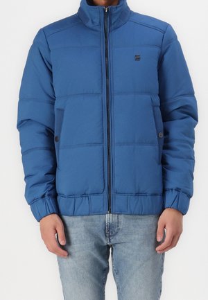 Homme portant une veste matelassée bleue avec une fermeture éclair à l'avant et un jean bleu, debout devant un fond blanc uni.