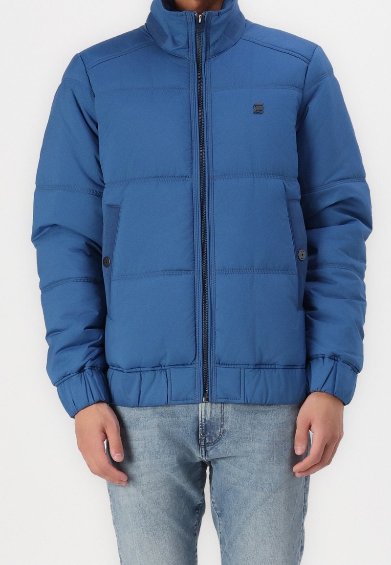 Homme portant une veste matelassée bleue avec une fermeture éclair à l'avant et un jean bleu, debout devant un fond blanc uni.
