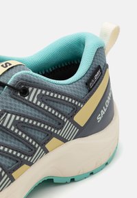 Grey and turquoise athletic shoe with textured fabric, striped accents, and a cushioned sole; features a pull tab and waterproof label.