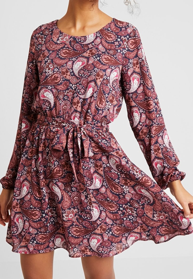 Femme portant une robe à manches longues, longueur genou, avec un motif cachemire rose et marron et une ceinture nouée à la taille.