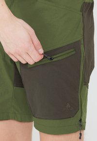 Gröna shorts med en mörkare grön sidoficka, dragkedjestängning och snörning. Tillverkade av ett lätt, texturerat tyg med sydda detaljer.