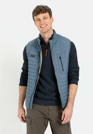 Light blue puffy vest with quilted texture, zippered front, and chest pocket, worn over a navy sweater and khaki cargo pants.