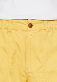 Gula byxor med elastisk midjeband och brun knapp, bärs tillsammans med en vit ribbad topp som syns vid midjan.