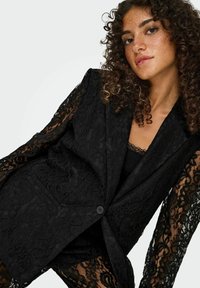 Črni blazer iz čipke z dolgimi prozornimi rokavi, enojnim zapiranjem na gumb in teksturiranim vzorcem tkanine z cvetličnimi motivi.
