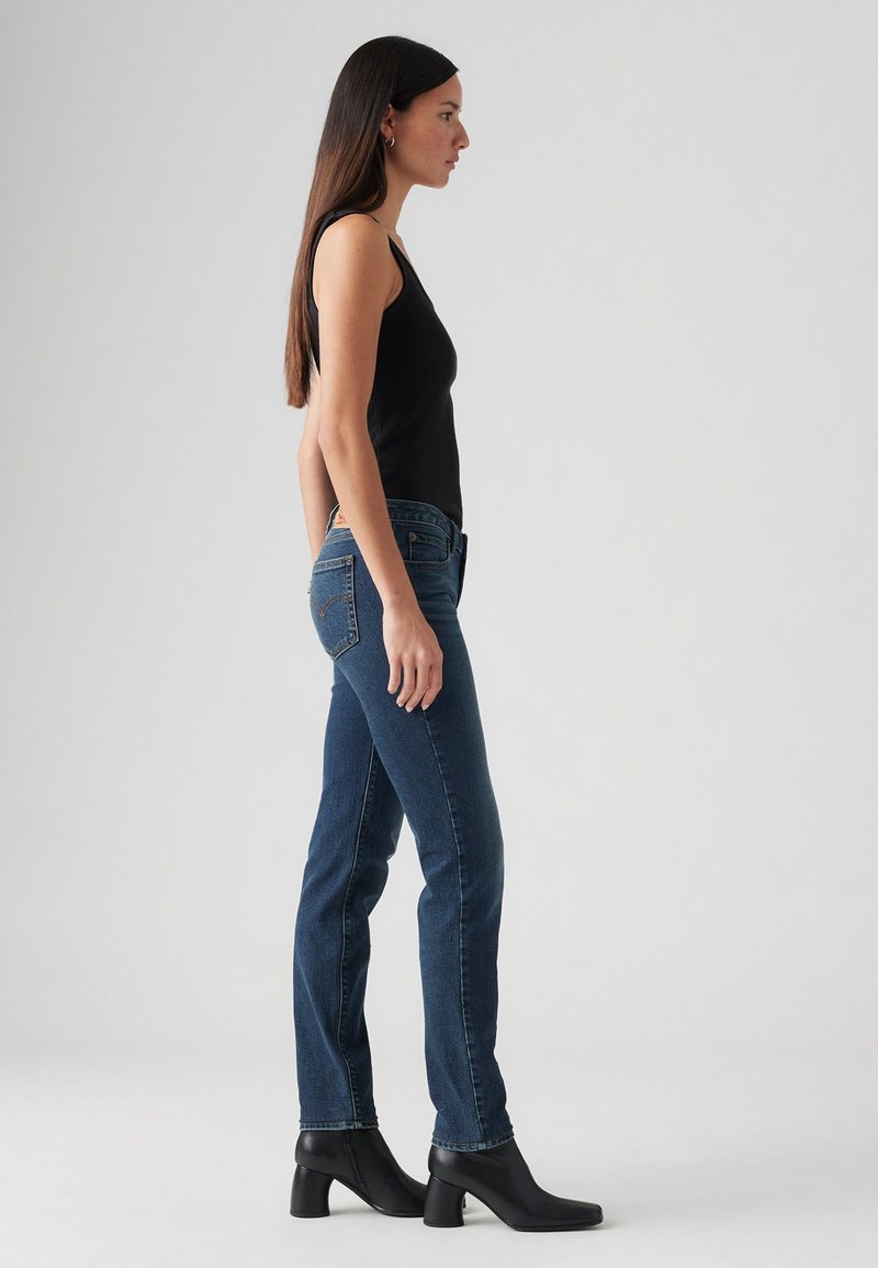 Jeans blu slim fit con vita alta, abbinati a un top senza maniche nero. Modella che indossa stivali neri con tacco, in piedi contro uno sfondo neutro.