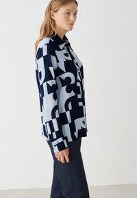 Marineblaues und hellblaues gemustertes Blusenshirt mit großen abstrakten Formen, einem klassischen Kragen und einer Knopfleiste, gefertigt aus einem glatten Stoff.