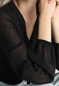 Blouse noire transparente avec manches bouffantes et encolure en V. Présente un motif à pois texturé et des poignets élastiques. Tissu lisse et léger.