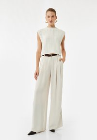 Strikket crop top i off-white med struktureret mønster, parret med lette, brede bukser i cremefarve, sorte spidse sko og et mørkt bælte.