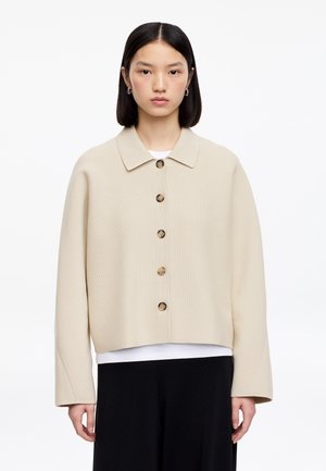 Kvinde med glat sort hår, iført en beige jakke med knapper over en hvid top og sorte bukser, stående mod en hvid baggrund.