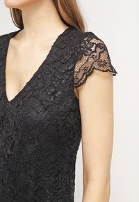 Kvinna i en svart spetsklänning med V-ringning och korta, genomskinliga spetsärmar mot en enfärgad bakgrund.