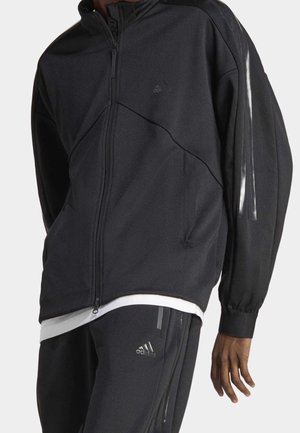 Homme portant une veste zippée noire Adidas et un pantalon assorti avec des logos, un sous-vêtement blanc visible à la taille, debout devant un fond uni.