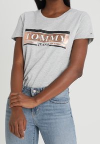 Camiseta de algodón gris con mangas cortas, que presenta un logo en negro y dorado rosado de "TOMMY JEANS" en el pecho. Combinada con unos jeans de mezclilla azul.