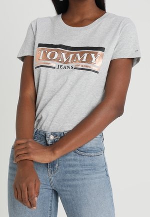 Camiseta de algodón gris con mangas cortas, que presenta un logo en negro y dorado rosado de "TOMMY JEANS" en el pecho. Combinada con unos jeans de mezclilla azul.