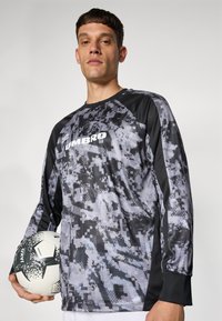 Langärmliges Fußballtrikot im grauen Camouflage-Muster mit schwarzen Akzenten. Besitzt ein Logo auf der Brust und gerippte Bündchen. Der Spieler hält einen Fußball.