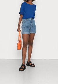 Top bluetto a maniche corte con dettagli in pizzo, pantaloni shorts in denim con risvolto, sandali neri e una borsa a tracolla arancione.